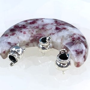 May include: Three silver cauldron charms with a loop for hanging. The charms are sitting on a crescent-shaped piece of pink and white speckled stone.