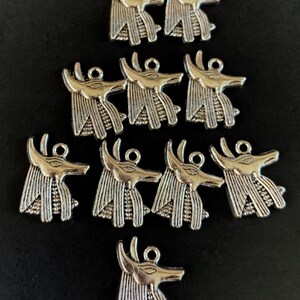 May include: Twelve silver charms in the shape of an Egyptian god's head with a human face and a jackal's head. The charms have a detailed design and are perfect for jewelry making.
