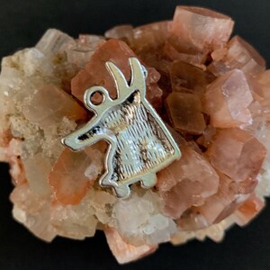 May include: A silver-colored metal charm shaped like a stylized deer with a textured surface, resting on a cluster of pink and white crystals.