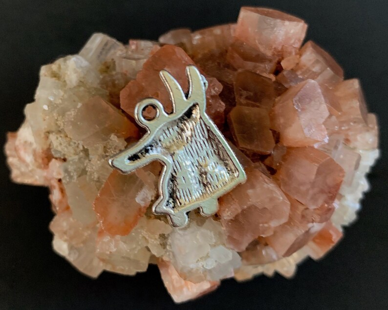 May include: A silver-colored metal charm shaped like a stylized deer with a textured surface, resting on a cluster of pink and white crystals.