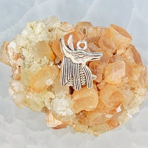 May include: A silver charm in the shape of an Egyptian god Anubis, with a detailed design, resting on a cluster of orange and white crystals.