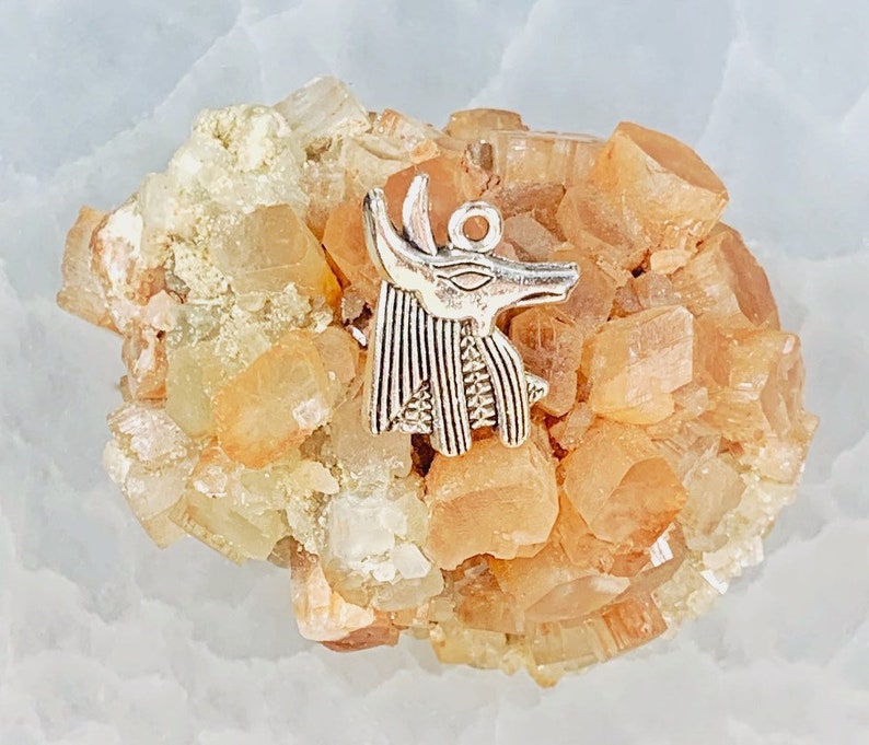 May include: A silver charm in the shape of an Egyptian god Anubis, with a detailed design, resting on a cluster of orange and white crystals.
