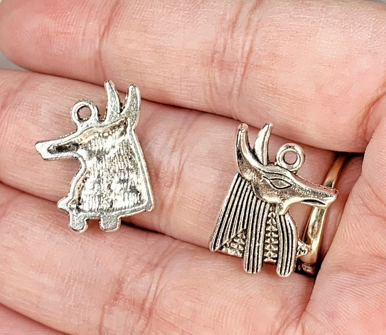 May include: Two silver charm pendants featuring an Egyptian Anubis jackal head design. One pendant is a simple profile, while the other is a more detailed depiction with a headdress.