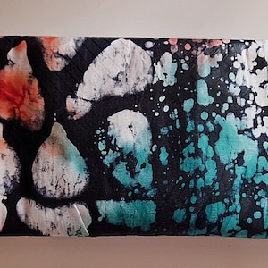 Puede incluir: Un artículo de tela rectangular con fondo negro y patrones abstractos en blanco, turquesa y naranja. El diseño tiene un aspecto tipo batik, con formas irregulares y salpicaduras de color. El artículo está doblado, lo que sugiere que podría ser una bufanda o un accesorio similar.