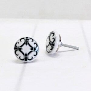 May include: Two white ceramic cabinet knobs with a black and gray decorative design. The knobs have a round shape and a metal screw for installation. The design features a symmetrical pattern. The knobs are suitable for drawers and cabinets.