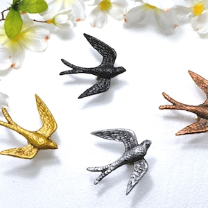 May include: Four metal swallow figurines in gold, black, silver, and copper colors are displayed on a white surface. The birds are in flight, with outstretched wings. White flowers and green leaves are in the background.