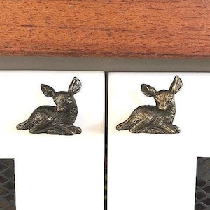 May include: Two bronze-colored cabinet knobs, each shaped like a resting fawn. The knobs are mounted on white cabinet doors, with a dark wood surface above. The fawns are detailed with visible ears and legs.