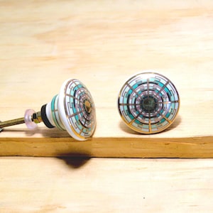 May include: Two decorative cabinet knobs with a geometric design in teal, gold, and white. The knobs are made of ceramic and have a brass screw and nut for installation. The knobs are on a wooden surface.