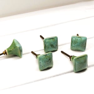 May include: A collection of five ceramic cabinet knobs. One knob is a light green, bell-shaped design, while the other four are square with a mottled turquoise finish. Each knob has a metal screw for installation.