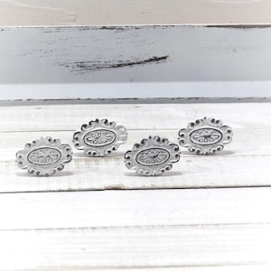 May include: Four white oval cabinet knobs with a floral design. The knobs are made of metal and have a distressed finish. The knobs are arranged in a row on a white wooden surface.