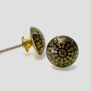 May include: Two decorative cabinet knobs with a black and gold floral design. The knobs have a round shape with a detailed pattern. The hardware includes a gold-colored base and a silver screw for installation.