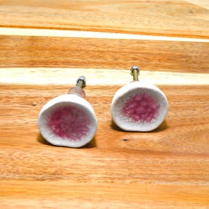 May include: Two white ceramic cabinet knobs with a pink, crystal-like interior. The knobs have a round shape and are mounted on metal hardware. The knobs are set against a wooden background.