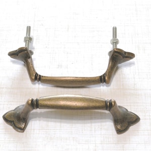 May include: Two antique bronze-colored drawer pulls with decorative ends and a curved handle. Each pull has a threaded bolt and nut for mounting. The hardware is designed for furniture restoration or DIY projects.