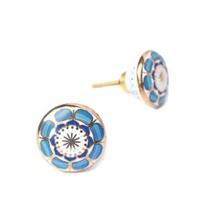 May include: Two decorative cabinet knobs with a floral design. The knobs feature a gold rim, a white center with a black starburst pattern, and blue petal-shaped accents. One knob is in focus, while the other is slightly blurred.
