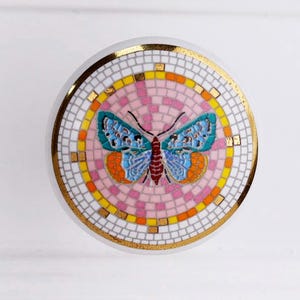 May include: A round, white ceramic cabinet knob with a mosaic butterfly design. The butterfly has blue, orange, and red details, set against a pink and white background. The knob is trimmed with gold.