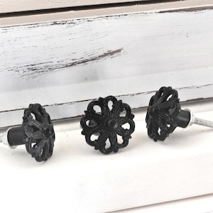 May include: Three black decorative cabinet knobs with a floral design. The knobs have a matte finish and a silver screw. The knobs are arranged on a white surface with a distressed wooden background.