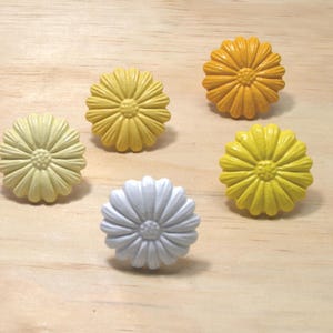 May include: Five daisy-shaped cabinet knobs in shades of yellow, white, and orange. The knobs are arranged on a light-colored wooden surface. Each knob has a detailed petal design and a central circular pattern.