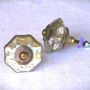 May include: Two decorative cabinet knobs. One is a starburst shape with a brass center and a distressed silver finish. The other is a faceted, clear glass knob with brass accents. Hardware includes a bolt, washer, and nut.