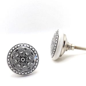 May include: Two white ceramic cabinet knobs with a black floral design. One knob is shown face-on, and the other is at an angle, revealing the metal screw and hardware. The knobs have a decorative, patterned design.
