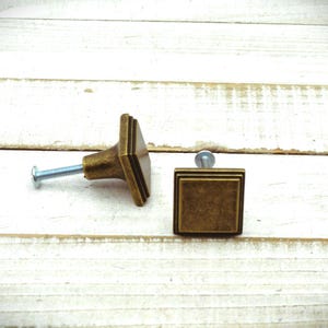 May include: Two antique brass square cabinet knobs with screws, set against a white wooden background. The knobs have a layered square design, showcasing a vintage aesthetic. Ideal for furniture restoration or adding a classic touch to any decor.
