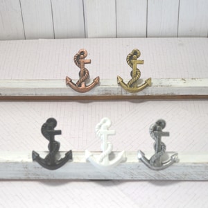 May include: A collection of six anchor-shaped cabinet knobs in various metallic finishes. The knobs include bronze, gold, black, white, and silver. Each anchor features a rope detail. The knobs are mounted on a white and brown wooden surface.