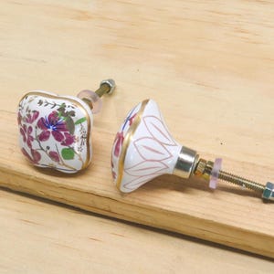 May include: Two decorative ceramic cabinet knobs with floral designs. One knob is square, the other is rounded. Both have white backgrounds with purple and gold accents. The knobs are on a wooden surface.