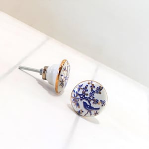May include: Two ceramic cabinet knobs with a white base, blue floral and bird design, and gold trim. One knob is attached to a silver screw, and the other is lying flat. The knobs are on a white surface.