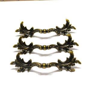May include: Three matching drawer pulls made of antique brass-toned metal. Each handle features a central circular knob flanked by symmetrical, ornate leaf-like scrollwork extending outward on both sides, creating a decorative horizontal shape.