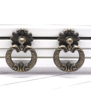 May include: Two antique bronze drawer pulls with ornate detailing. Each pull features a decorative backplate and a ring-shaped handle. The hardware is mounted on a white surface, creating a contrast.
