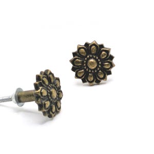 May include: Two antique brass-colored flower-shaped cabinet knobs on a white background. One knob is lying on its side, showing the screw, and the other is upright. The knobs have a detailed floral design.