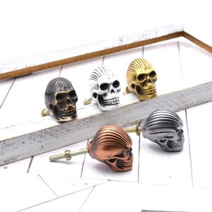 May include: Five decorative skull cabinet knobs in various metallic finishes: black, white, gold, copper, and silver. Each skull knob features a detailed design and is mounted on a screw for easy installation. Perfect for adding a unique touch to furniture.