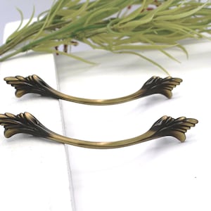 May include: Two curved metal drawer pulls with an antique brass finish. Each handle features a central arch and decorative, wing-like ends with detailed ridges. The hardware has a smooth, metallic texture with darker shading in the recessed areas.