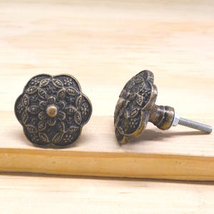May include: Two antique bronze cabinet knobs with a floral design. The knobs have a detailed, embossed pattern and a threaded screw for installation. The knobs are displayed on a light wood surface.