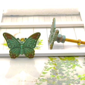 May include: Two decorative butterfly cabinet knobs. The knobs are teal and brown with a detailed butterfly design. The hardware includes a gold-colored bolt and nut. The knobs are displayed on a white frame.
