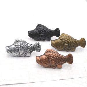 May include: Four decorative fish figurines in varying metallic finishes: silver, bronze, copper, and black. Each fish has a textured scale pattern and is positioned on a white surface. These small sculptures could be used as cabinet knobs or decorative accents.