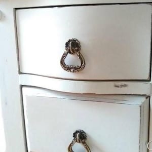 May include: Close-up of a white cabinet with two drawers. Each drawer has an ornate, antique-style brass pull. The cabinet has a distressed finish, with visible wear and tear on the edges, giving it a vintage appearance.