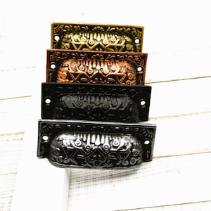 May include: A stack of four decorative drawer pulls in various metallic finishes: gold, copper, black, and silver. Each pull features an ornate, embossed design with a curved handle and two screw holes for mounting. The pulls are rectangular and have a vintage aesthetic.