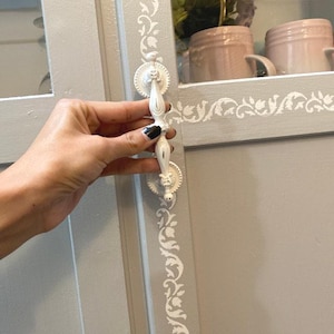 May include: A white, ornate cabinet pull with a decorative design. The pull is held against a gray cabinet door with white floral accents. The cabinet door is part of a larger cabinet with a glass door.