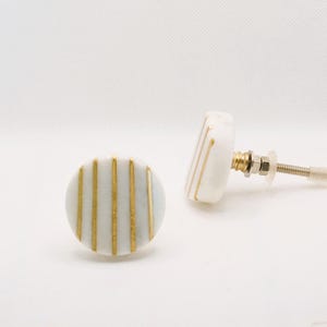 May include: Two white and gold cabinet knobs. Each knob is round with vertical gold stripes. One knob is lying flat, while the other is upright, showing the screw and hardware.