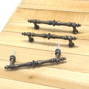 May include: Three antique bronze cabinet pulls with decorative detailing. The pulls are arranged on a light wood surface, showcasing their ornate design and vintage aesthetic. Each pull has a bar shape with decorative ends and mounting hardware.