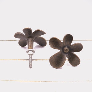 May include: Two bronze-colored flower-shaped cabinet knobs. One knob is attached to a screw and washer, while the other is separate. The knobs have a vintage, antique appearance and are set against a white background.