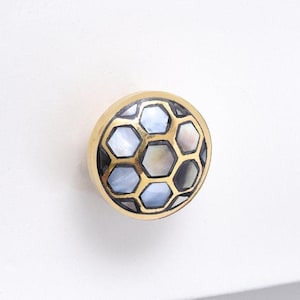 May include: A round cabinet knob with a gold-colored rim and a hexagonal pattern. The pattern is filled with iridescent, pearlescent shell pieces. The knob is mounted on a white surface.