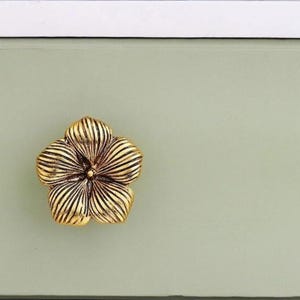 May include: Close-up of a gold-colored, flower-shaped drawer pull. The drawer pull is mounted on a light green surface. The flower has detailed, textured petals. The background is a light green color.