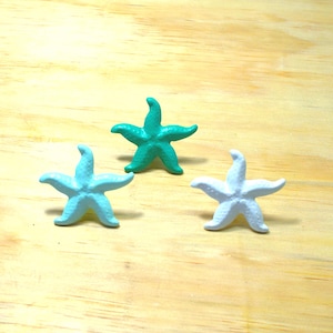 May include: Three small starfish figurines in shades of light blue, teal, and white are arranged on a light brown wooden surface. The starfish have five arms and a textured appearance, suggesting a coastal or nautical theme.