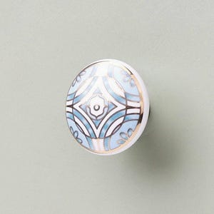 May include: A round ceramic cabinet knob featuring a white base with a symmetrical geometric pattern in shades of light blue and dark blue, accented by thin gold metallic lines that define the intricate shapes across the surface of the knob.