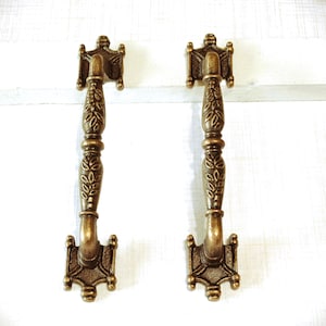 May include: Two antique bronze cabinet pulls. Each pull features a decorative handle with a floral design and ornate end pieces. The pulls are mounted on a white surface.