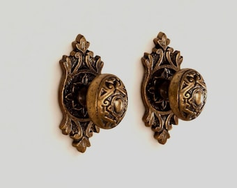 May include: Two matching antique brass door knobs featuring intricate embossed scrollwork and floral patterns on both the circular handle and the elongated, decorative backplate, showcasing a weathered, dark metallic finish.