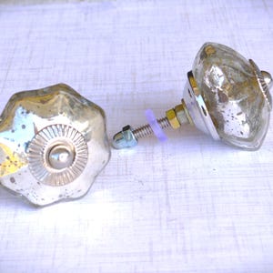 May include: Two antique-style cabinet knobs with a distressed, mercury glass finish. The knobs have a starburst design and are attached to metal hardware with a screw and a purple rubber washer. These decorative knobs are ideal for furniture restoration.