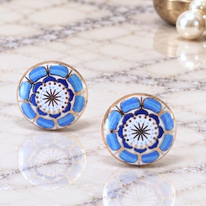 May include: Two round stud earrings featuring a central white circular design with a black starburst pattern, surrounded by a ring of blue petals and gold-toned metal edges. The earrings have a smooth, glossy finish and a symmetrical floral-inspired appearance.