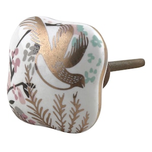 May include: A white ceramic cabinet knob with a gold bird design. The knob features a square shape with a rounded edge and a threaded metal rod for installation. The bird is depicted in flight, surrounded by floral and botanical accents in pink, gray, and green.